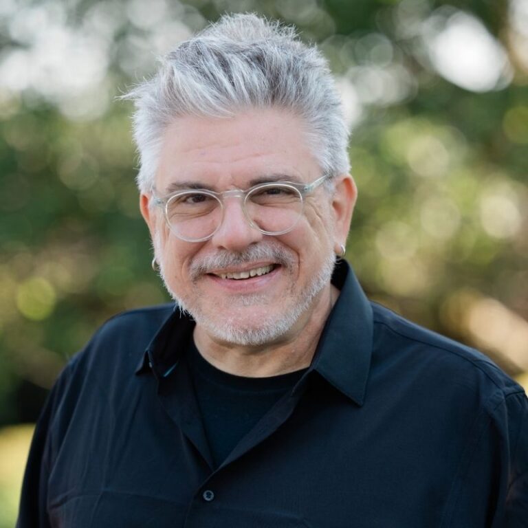 A man with silver-gray hair swept up and back, a matching mustache and beard, and clear wire-frame glasses smiles broadly outdoors against a softly blurred green background with small stud earrings visible. He wears a black button-down shirt layered over a black t-shirt.