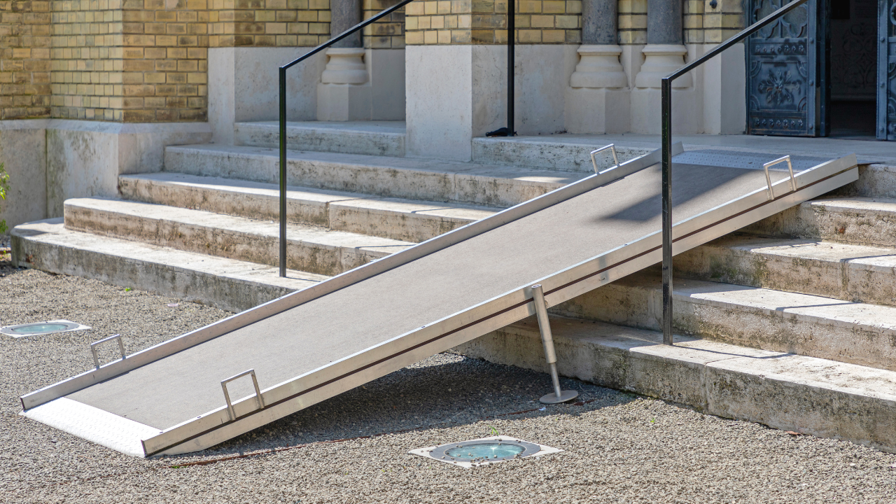 A portable metal accessibility ramp with handrails is placed over stone steps leading to a brick building entrance, providing wheelchair access to the doorway.