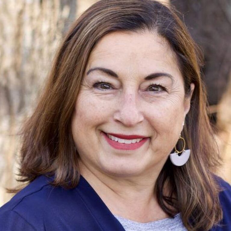 A woman with shoulder-length brown hair smiles broadly, wearing bold red lipstick, against a blurred outdoor background of tree bark and foliage. She wears a navy cardigan over a heathered gray top, with gold hoop earrings featuring a white geometric charm.