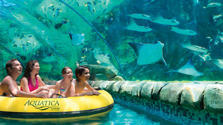 A family rides in a yellow inflatable raft through a water park attraction with a clear underwater tunnel, watching fish and rays swim overhead.