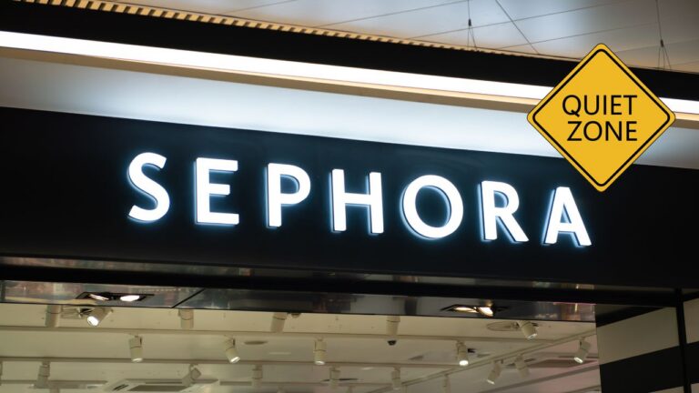 A yellow diamond-shaped Quiet Zone caution sign hangs in the upper right corner above a storefront, with a black-and-white illuminated Sephora sign spanning the entry above. The store entrance below shows track lighting, glass display cases, and a glimpse of the softly lit interior.