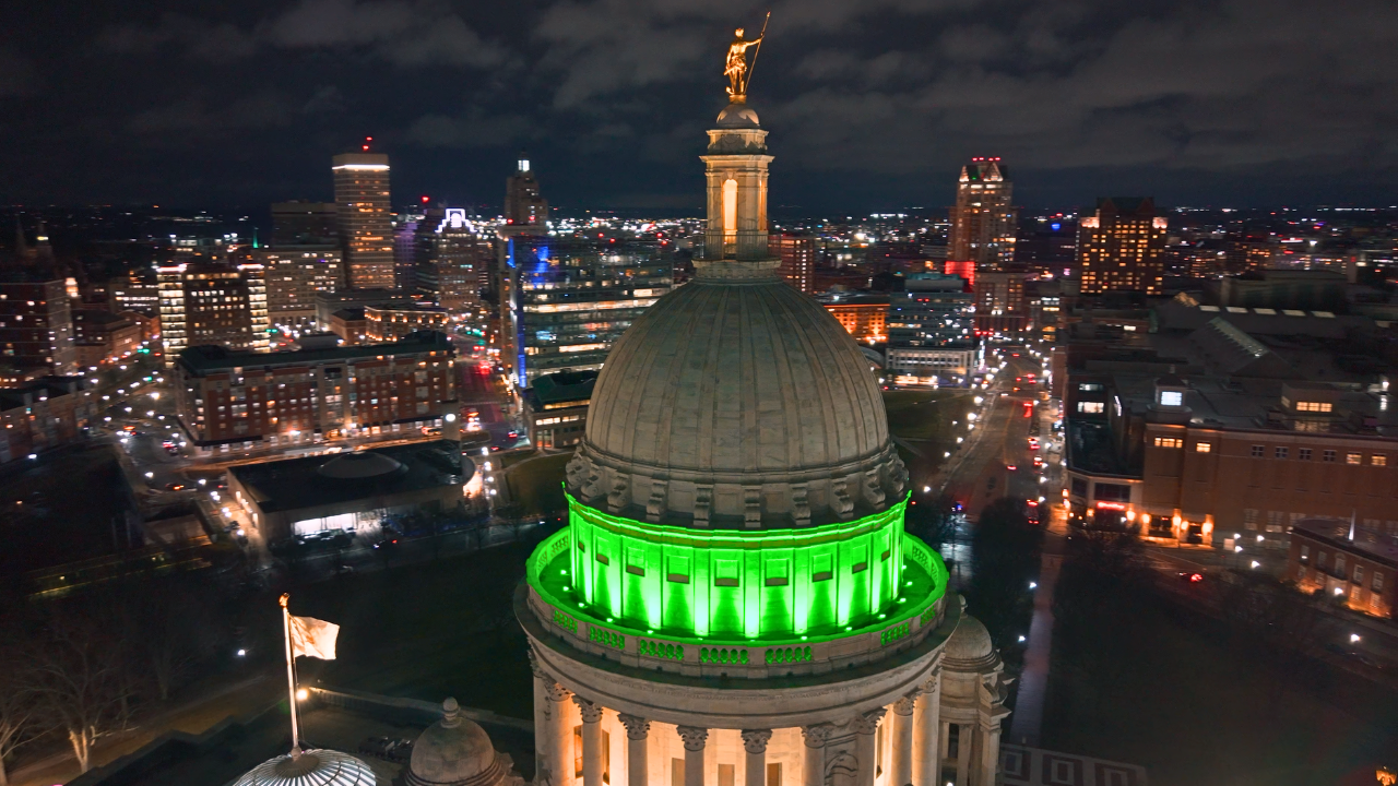 Aerial nighttime view of the Rhode Island State House dome lit bright green, with the gold statue on top visible against a city skyline filled with lights and buildings in Providence.