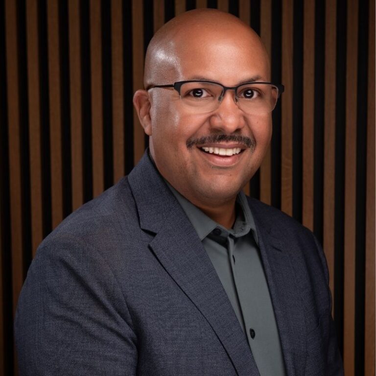 A bald man with a mustache and thin-framed glasses smiles broadly against a vertical wood slat background. He wears a dark gray blazer over a green button-down shirt.