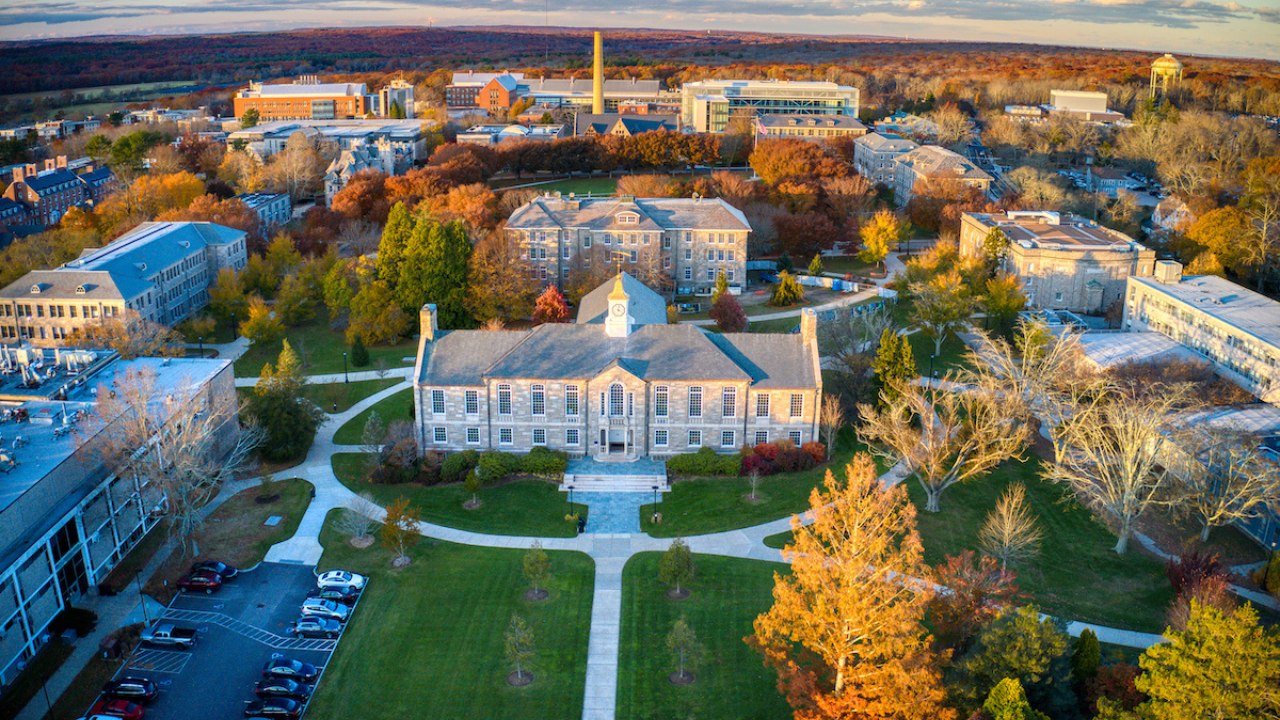 Aerial view of a college campus in autumn, centered on a symmetrical historic stone building with a small clock tower and columned entrance. Tree-lined walkways radiate outward across green lawns, surrounded by academic buildings. The landscape is filled with fall foliage in shades of orange, red, and yellow, with parking areas and a distant tree-covered horizon visible under a partly cloudy sky.