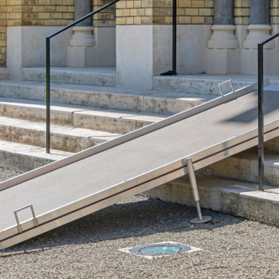 A portable metal accessibility ramp with handrails is placed over stone steps leading to a brick building entrance, providing wheelchair access to the doorway.