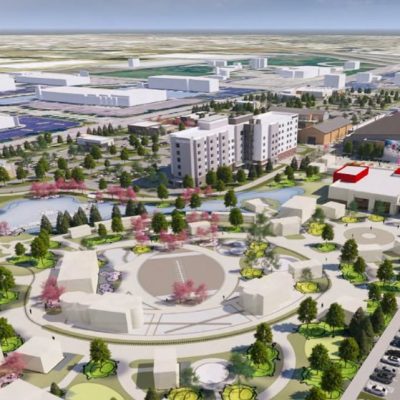 An aerial rendering shows a large, master-planned development from an elevated perspective. At the center is a circular plaza with a wide, open gathering space and geometric walkways radiating outward. Surrounding the plaza are several low-rise buildings arranged in a ring-like pattern, connected by curved pedestrian paths and landscaped green spaces. Clusters of trees—some with pink blossoms—are scattered throughout the site, along with small lawns, planted beds, and winding sidewalks. A pond with a curved shoreline sits to the left of the central area, bordered by trees and walking paths. Behind the plaza, a mid-rise residential-style building with multiple floors stands prominently, with additional large buildings nearby that resemble event spaces, retail structures, or community facilities. One building features red rectangular accents on its roof. To the right side of the image is a long parking lot filled with rows of parked cars. In the distance, expansive industrial or warehouse buildings and large parking areas stretch toward the horizon, indicating the development is situated within a broader commercial or mixed-use district. The overall image appears to be a digital architectural visualization rather than a photograph.