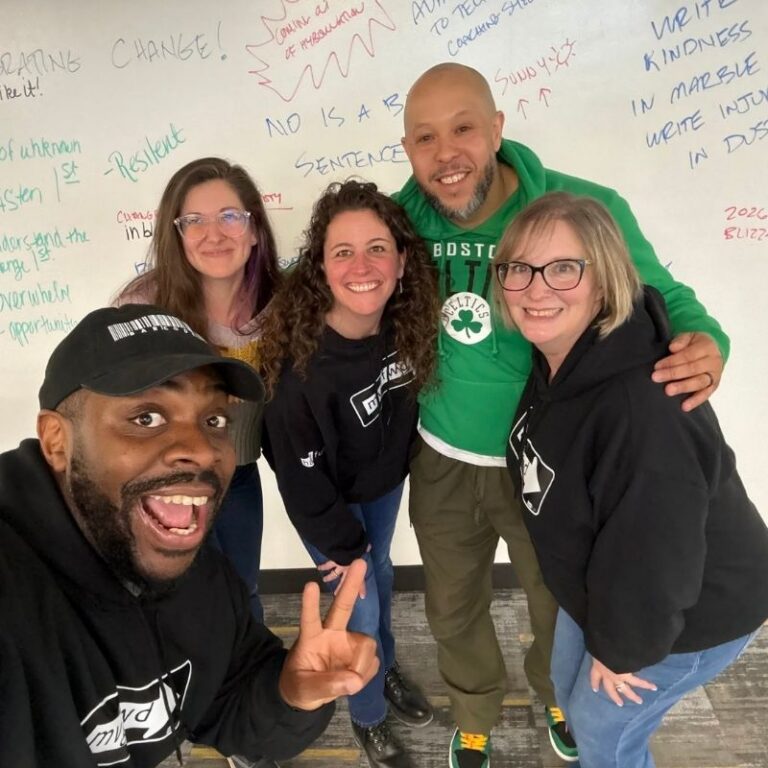 A group selfie of five people posing closely together and smiling, taken in front of a large whiteboard covered in colorful handwritten notes and phrases like Change and No Is A Complete Sentence. In front, a man wearing a black cap and matching branded hoodie leans in close to the camera making a peace sign, while behind him a bald man in a green Celtics hoodie has his arms around two women in matching black branded hoodies and another woman in glasses stands to the side, all smiling warmly at the camera.